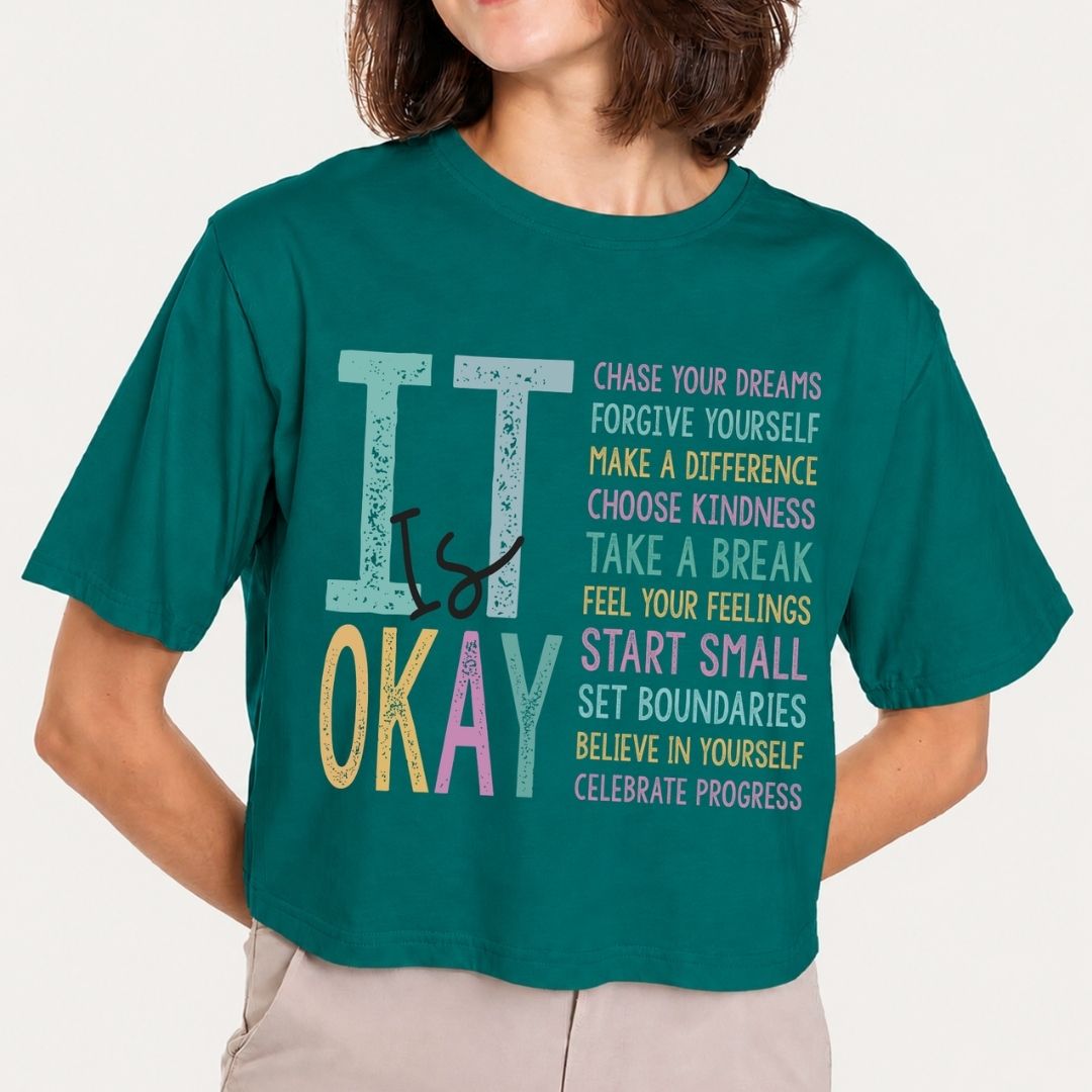 A woman wearing a teal green cropped t-shirt with a colorful graphic that reads 'IT IS OKAY' with motivational text on the front, paired with light beige pants, against a plain off-white background.