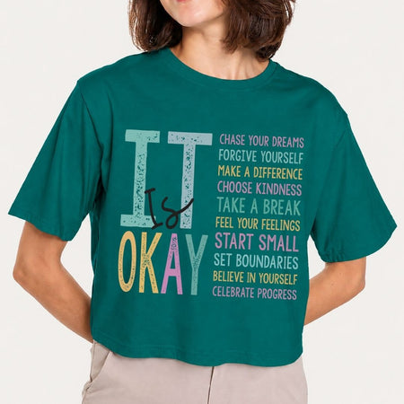 A woman wearing a teal green cropped t-shirt with a colorful graphic that reads 'IT IS OKAY' with motivational text on the front, paired with light beige pants, against a plain off-white background.