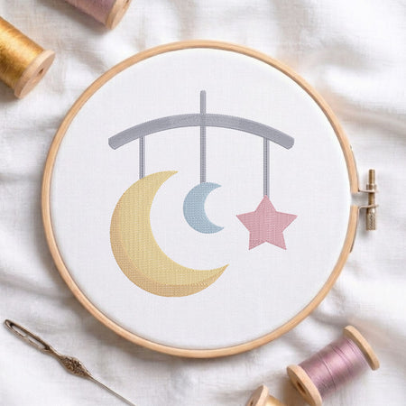 A circular embroidery hoop holding white fabric with a stitched mobile design: a large yellow crescent moon, a small light blue crescent moon, and a pink star under a gray arch. Spools of thread and a needle are arranged around it on a white fabric background.