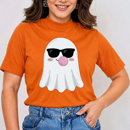 A woman wearing a bright orange short-sleeve t-shirt with a cartoon ghost graphic wearing black sunglasses and blowing a pink bubblegum bubble. She is also wearing blue jeans against a plain light gray background.