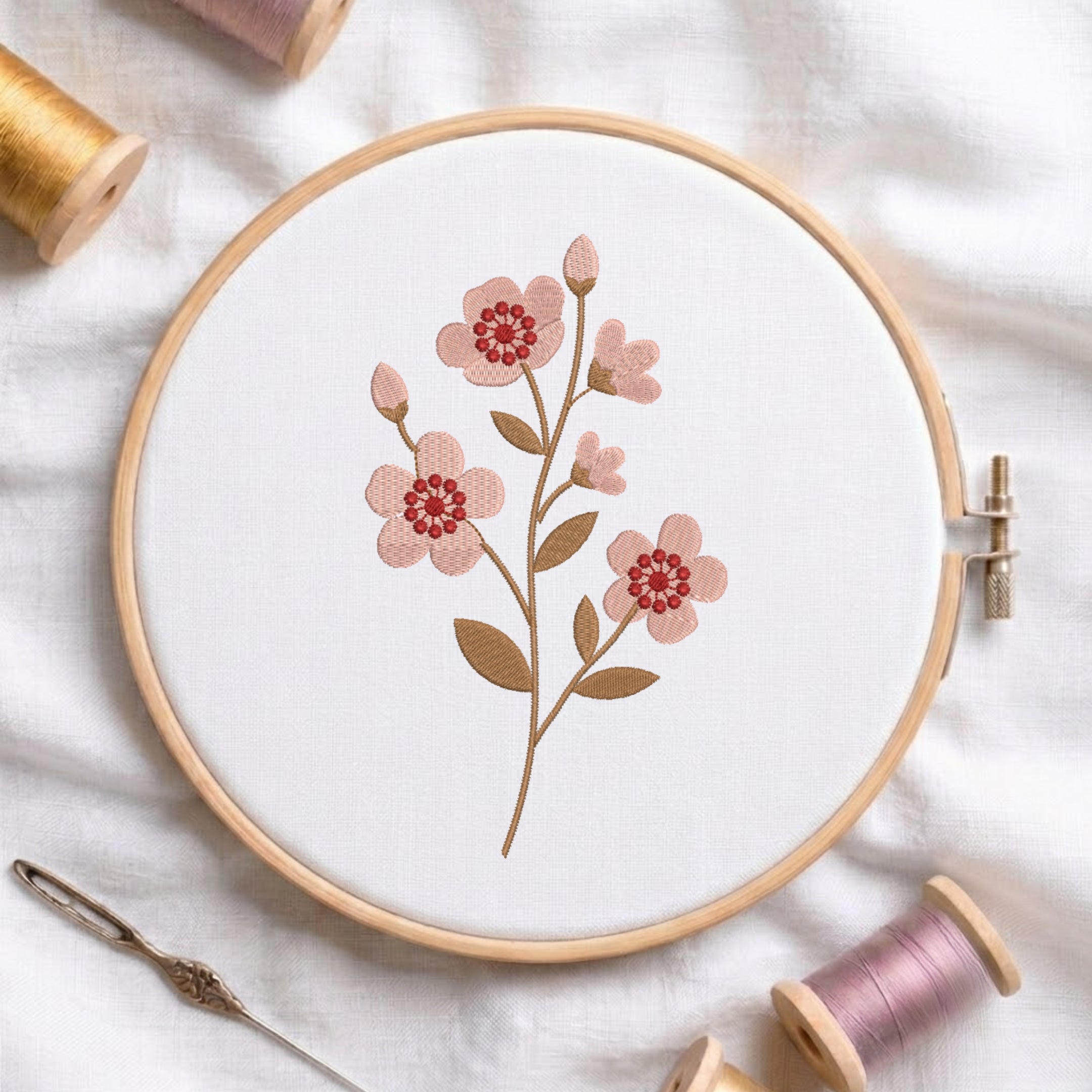 A wooden embroidery hoop holds white fabric with a floral design of pink blossoms and green-brown leaves. Spools of thread and a needle are arranged around it on a white fabric background.