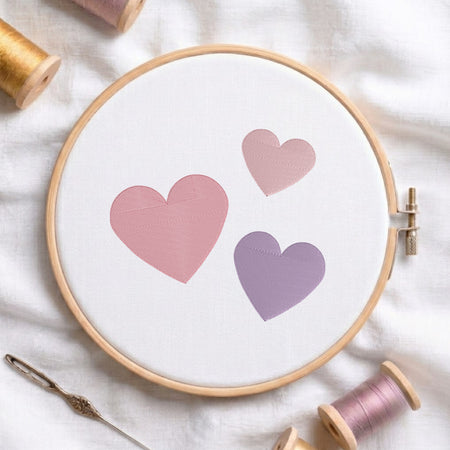 A round embroidery hoop holding white fabric with three stitched hearts in soft pink and lavender tones, styled on a white cloth with thread spools and a needle around it.