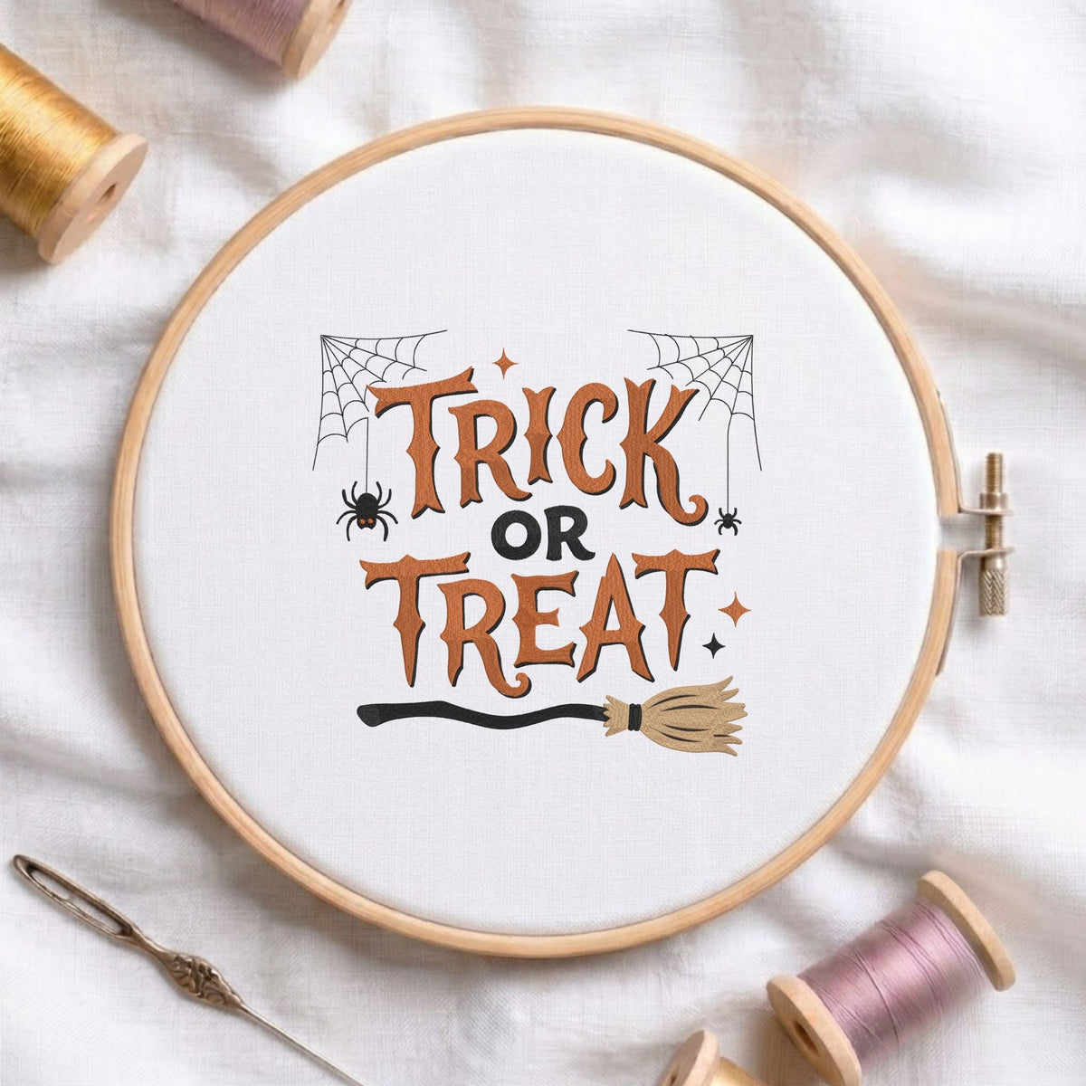 "Trick or Treat" lettering with spider webs, spiders, and a witch's broom machine embroidery design shown in a wooden embroidery hoop on white fabric with thread spools and a needle