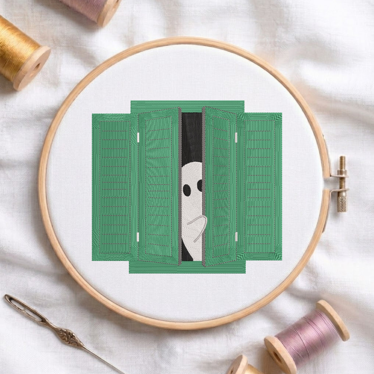 A circular embroidery hoop holds white fabric with a green shutters design and a peeking white face motif. The hoop is styled on a white fabric background with spools of thread and a needle visible around it.