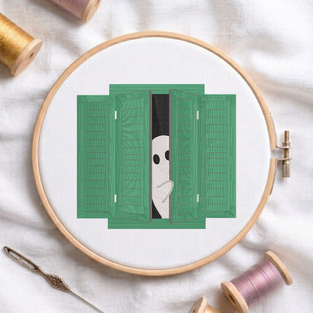 A circular embroidery hoop holds white fabric with a green shutters design and a peeking white face motif. The hoop is styled on a white fabric background with spools of thread and a needle visible around it.