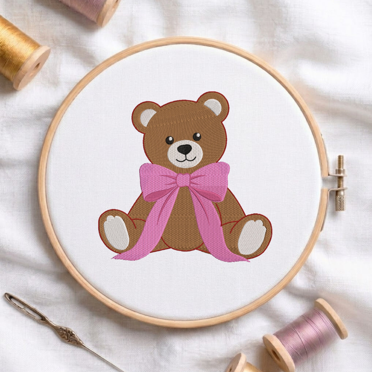 A circular embroidery hoop holding white fabric with a stitched brown teddy bear wearing a pink bow. The hoop is styled on a white fabric background with spools of thread and a needle around it.