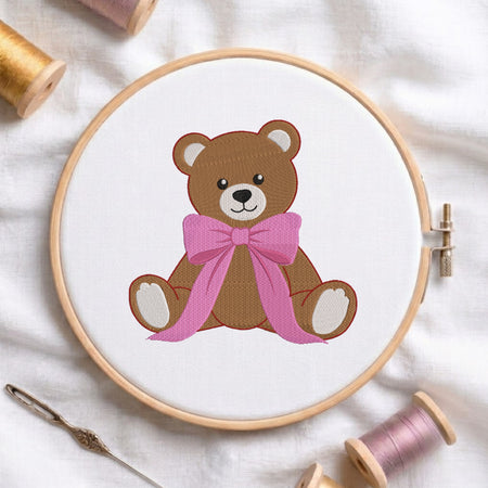 A circular embroidery hoop holding white fabric with a stitched brown teddy bear wearing a pink bow. The hoop is styled on a white fabric background with spools of thread and a needle around it.