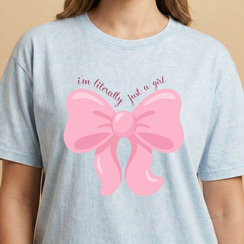 Person wearing a light blue tie-dye style t-shirt featuring a large pink ribbon bow design with the text "i'm literally just a girl".