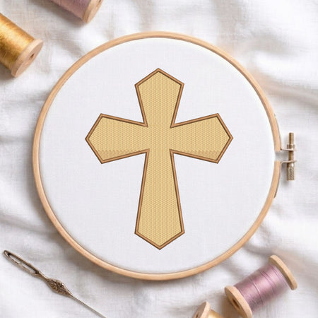 An embroidery hoop on a white cloth displays a finished embroidery design of a textured gold-yellow Christian cross with pointed ends and a brown outline, flanked by spools of thread and a needle.