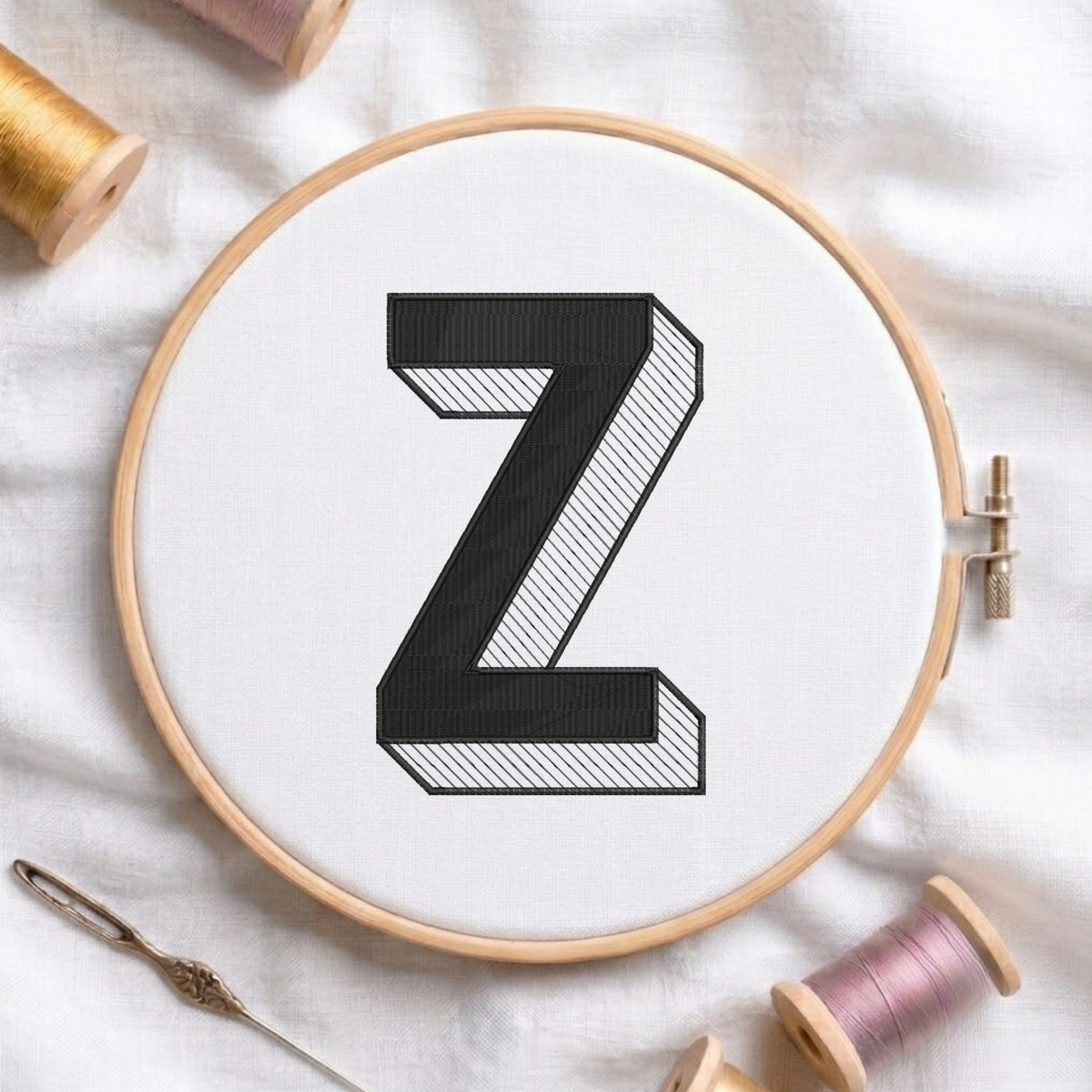 A wooden embroidery hoop holding white fabric with a large black letter Z, surrounded by spools of thread and a needle on a white fabric background.