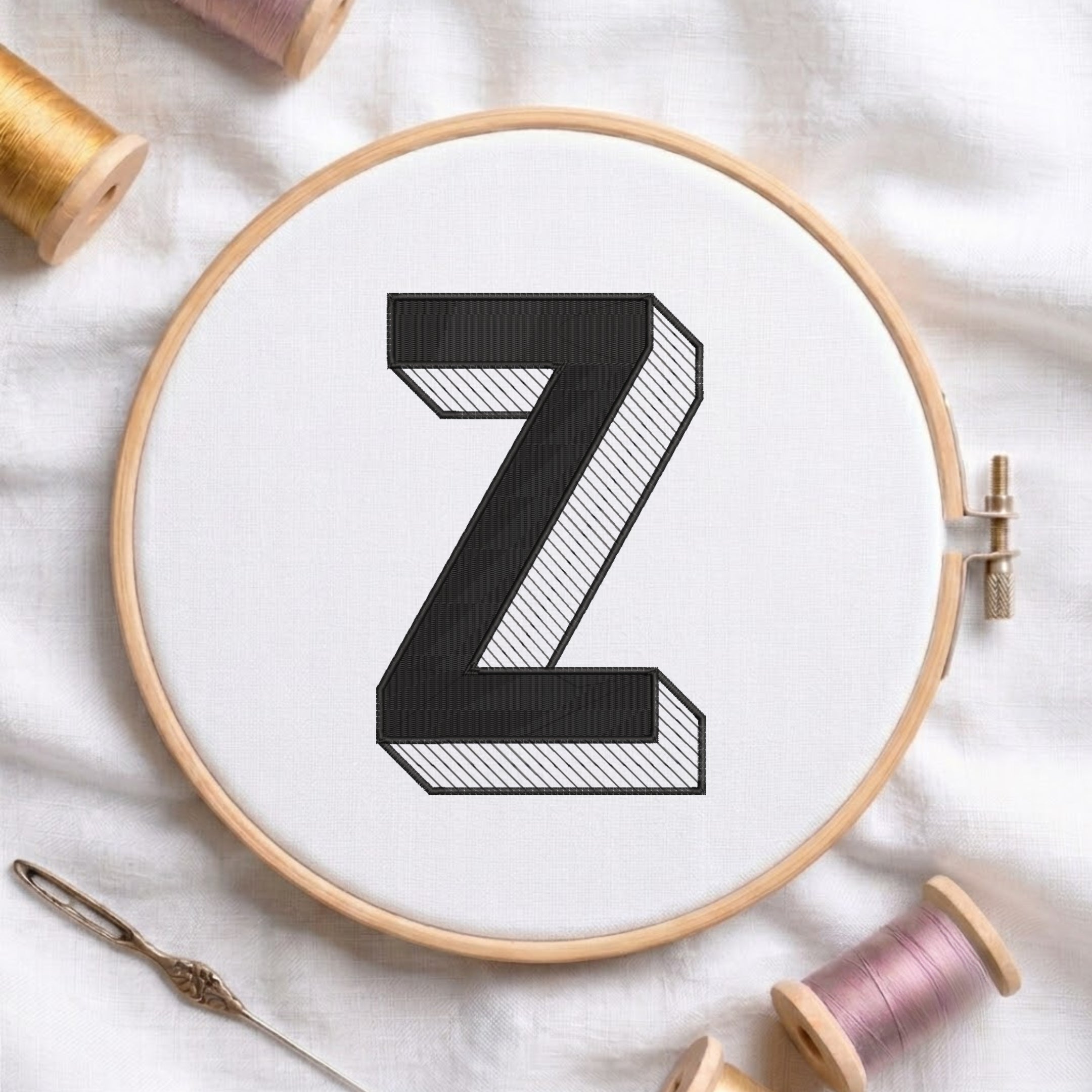 A wooden embroidery hoop holding white fabric with a large black letter Z, surrounded by spools of thread and a needle on a white fabric background.