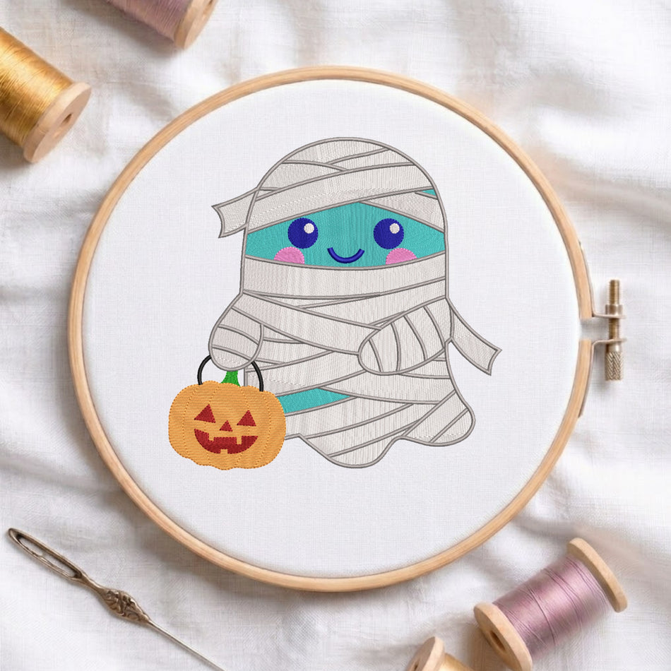 A wooden embroidery hoop displays a cute cartoon mummy ghost with blue eyes and a pumpkin bag, surrounded by spools of thread and a needle on a white fabric background.