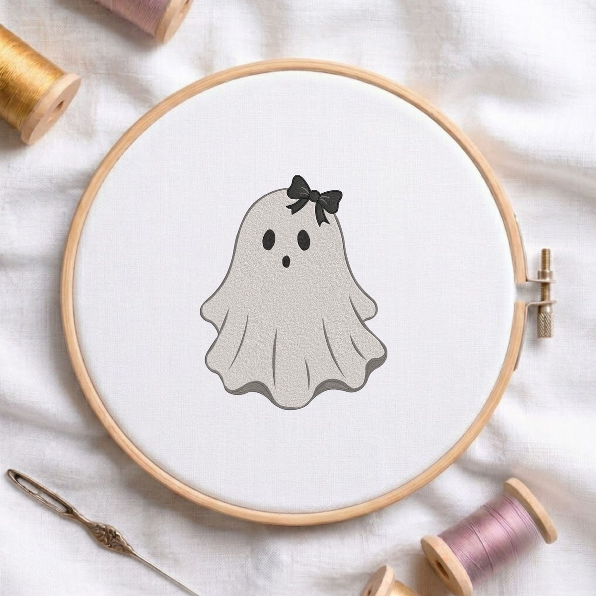 Cute ghost wearing a small black hair bow machine embroidery design shown in a wooden embroidery hoop on white fabric with thread spools and a needle   