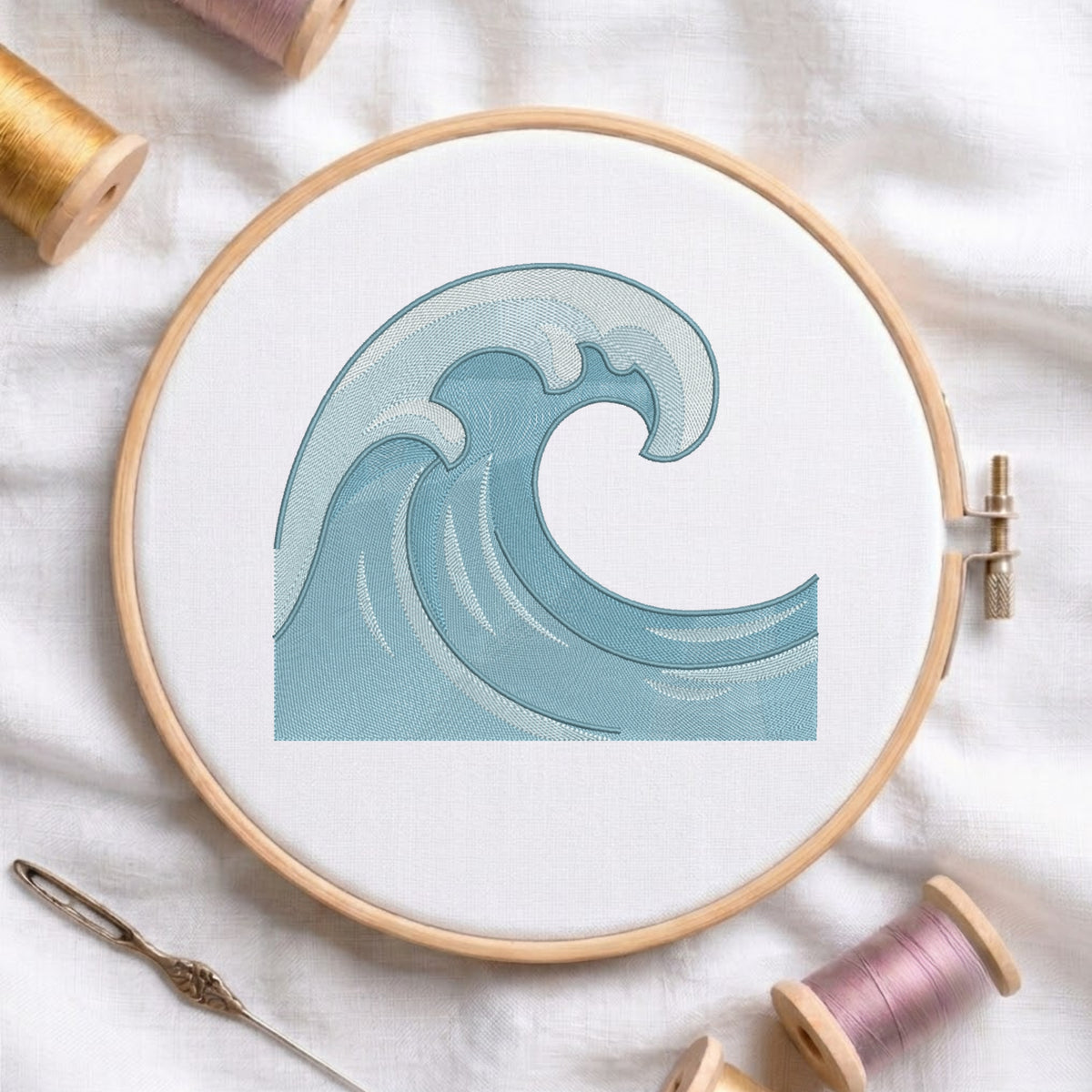 A wooden embroidery hoop holding white fabric with a stitched blue wave design, surrounded by spools of thread and a needle on a white fabric background.