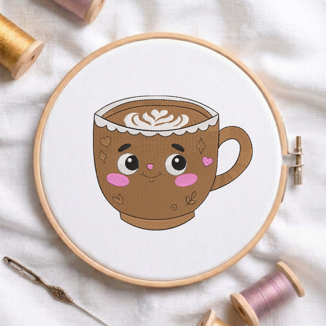 A wooden embroidery hoop holds white fabric with a cartoon-style coffee cup design in brown, cream, pink, and black. The hoop is styled on a white fabric background with spools of thread and a needle nearby.