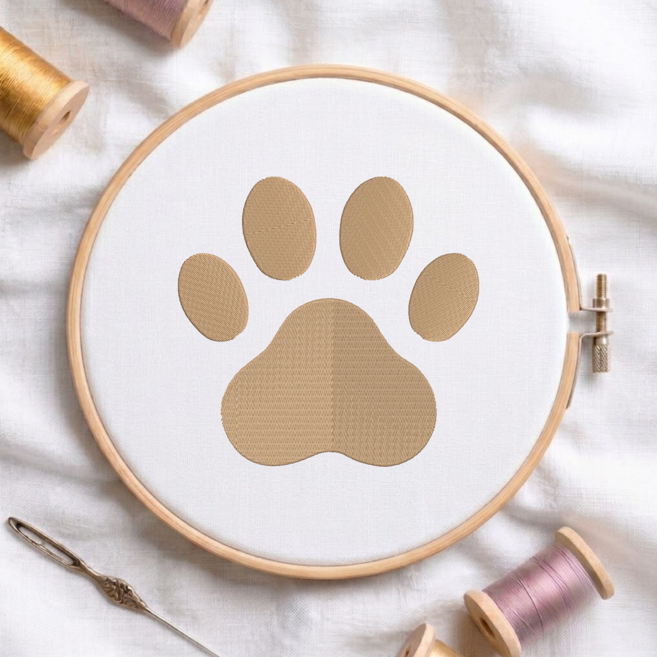 A circular embroidery hoop holding white fabric with a tan paw-print design, surrounded by spools of thread and a needle on a white fabric background.