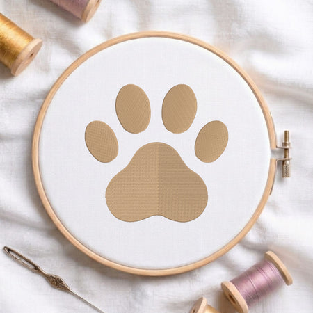 A circular embroidery hoop holding white fabric with a tan paw-print design, surrounded by spools of thread and a needle on a white fabric background.