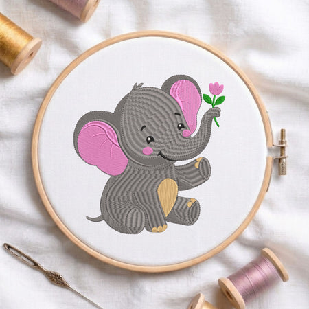 A circular embroidery hoop holding white fabric with a stitched cartoon elephant in gray and pink holding a small pink flower. The hoop is styled on a white fabric surface with spools of thread and a needle around it.