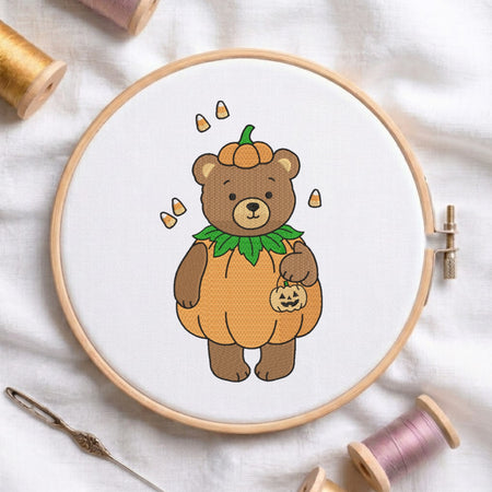 An embroidered brown bear wearing a pumpkin costume with candy corn accents, stitched inside a wooden hoop on white fabric with sewing threads.