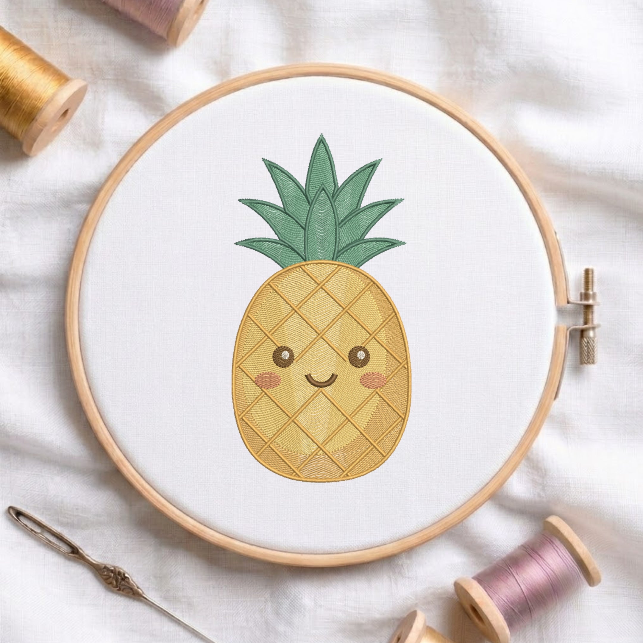 A wooden embroidery hoop holding white fabric with a cute embroidered pineapple design in yellow, green, and pink, surrounded by spools of thread and a needle on a white fabric background.
