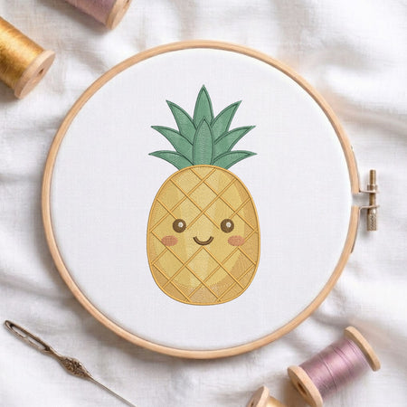 A wooden embroidery hoop holding white fabric with a cute embroidered pineapple design in yellow, green, and pink, surrounded by spools of thread and a needle on a white fabric background.
