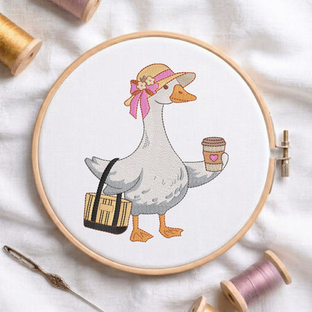 A cross-stitch embroidery design of a white goose wearing a tan hat with a pink bow and carrying a black-and-tan bag, shown inside a wooden embroidery hoop on a white fabric background with spools of thread and a needle nearby.