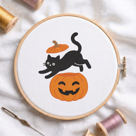 Playful black cat leaping over a carved jack-o-lantern pumpkin with its lid hovering above Halloween machine embroidery design shown in a wooden embroidery hoop on white fabric with thread spools and a needle
