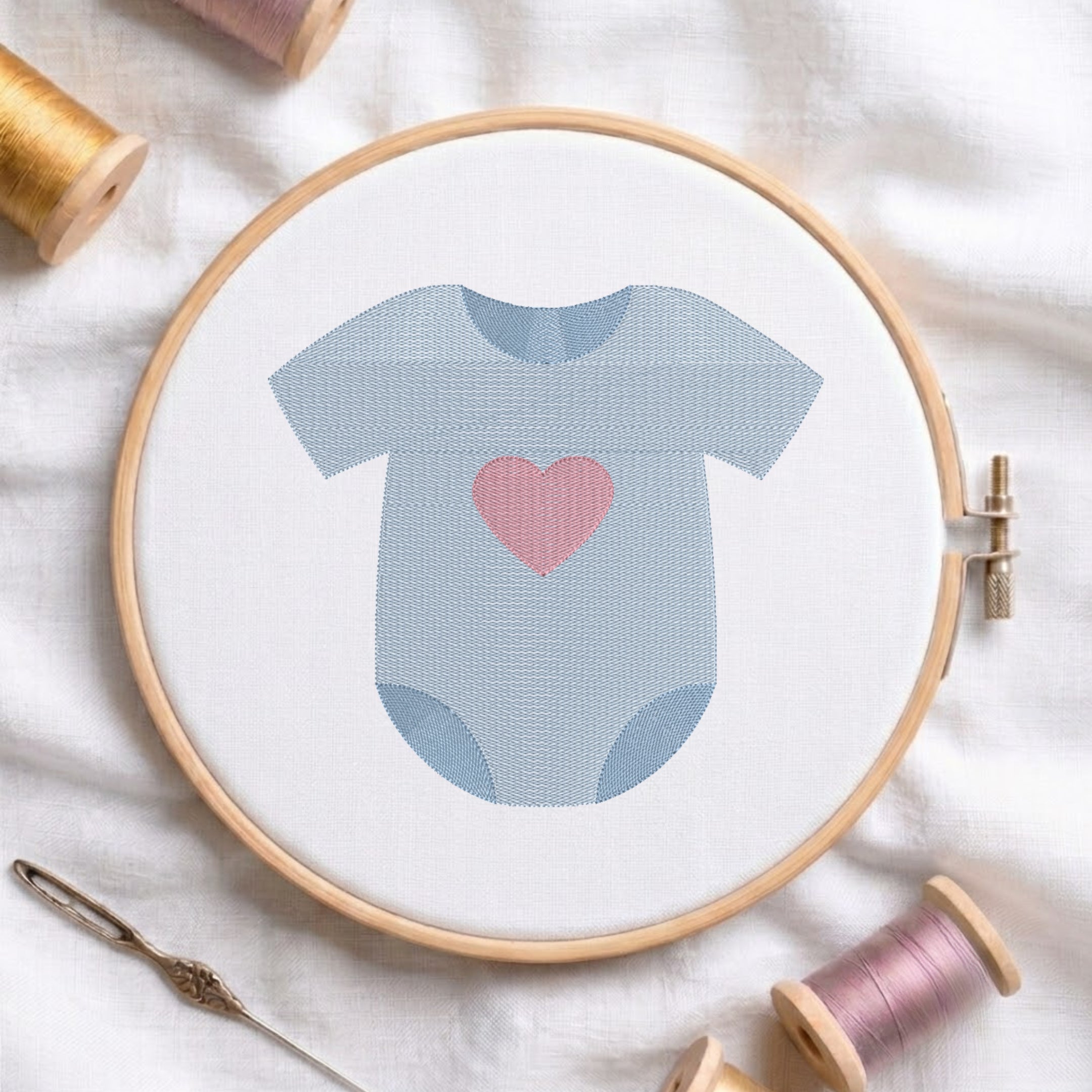 A baby bodysuit embroidery design held in a wooden hoop, featuring a light blue onesie with a pink heart on the chest. The hoop is styled on a white fabric background with spools of thread and a needle.