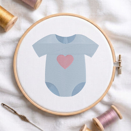 A baby bodysuit embroidery design held in a wooden hoop, featuring a light blue onesie with a pink heart on the chest. The hoop is styled on a white fabric background with spools of thread and a needle.