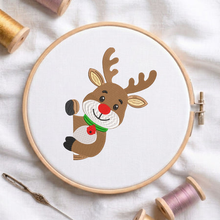 A circular embroidery hoop holding a white fabric panel with a cartoon reindeer design. The reindeer is brown with beige ears and antlers, a red nose and bell, and a green collar.
