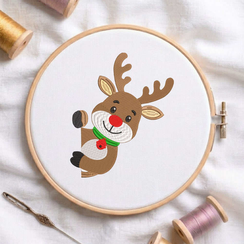 A circular embroidery hoop holding a white fabric panel with a cartoon reindeer design. The reindeer is brown with beige ears and antlers, a red nose and bell, and a green collar.