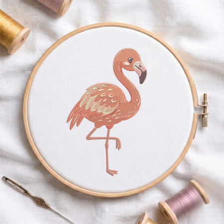A circular embroidery hoop holding white fabric with a stitched flamingo design in peach and brown tones. The hoop is styled on a white fabric surface with spools of thread, a needle, and a pin around it.