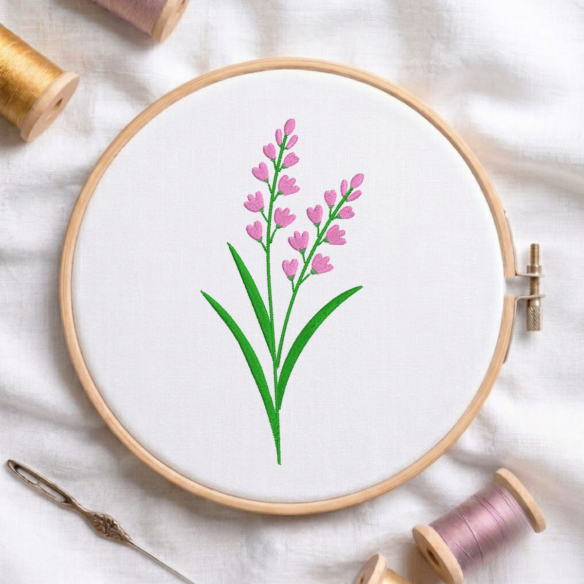 Pink lavender sprigs with green stems and leaves embroidered inside a round wooden hoop surrounded by thread spools.