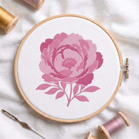 A wooden embroidery hoop holds white fabric with a large pink floral design stitched in the center. A needle and thread spools are arranged around it on a soft white fabric background.