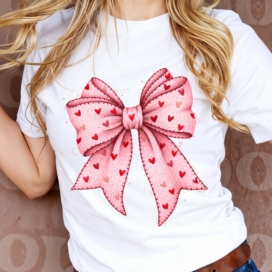 White t-shirt with a large pink bow design featuring red hearts on a patterned background.