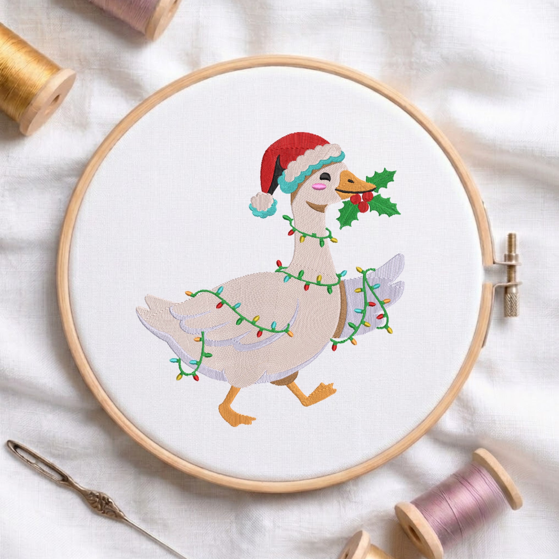 A circular embroidery hoop holds a white fabric with a stitched illustration of a goose wearing a red Santa hat and wrapped in multicolored Christmas lights, holding holly. The hoop is styled on white fabric with spools of thread and a needle around it.