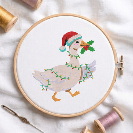 A circular embroidery hoop holds a white fabric with a stitched illustration of a goose wearing a red Santa hat and wrapped in multicolored Christmas lights, holding holly. The hoop is styled on white fabric with spools of thread and a needle around it.