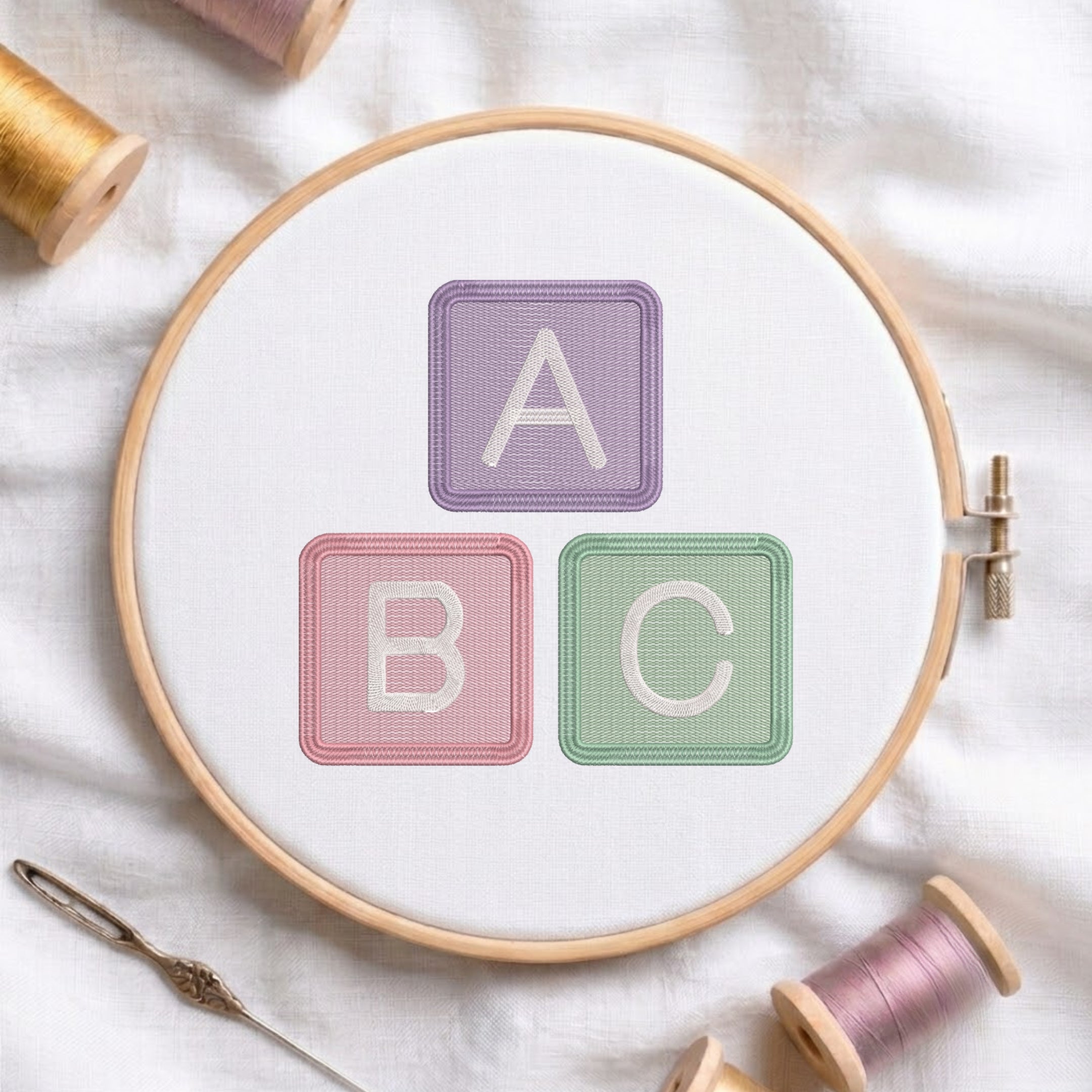 A wooden embroidery hoop on white fabric, showing three pastel embroidered letter blocks reading A, B, and C. Spools of thread, a needle, and a thimble are arranged around it.