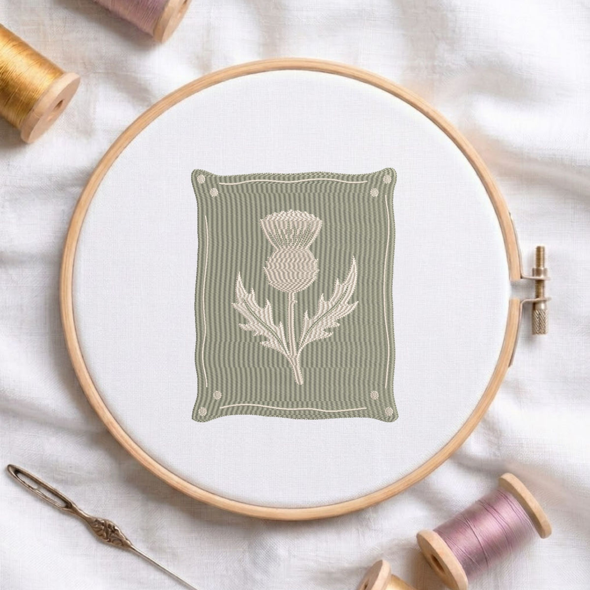 A circular embroidery hoop holds white fabric with a stitched design of a thistle flower on a muted sage green background. The hoop is styled on a white fabric surface with spools of thread and a needle around it.