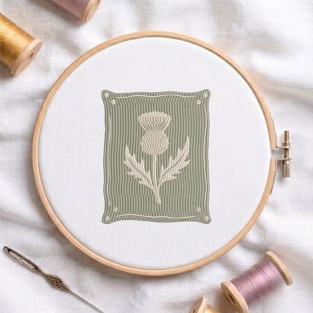 A circular embroidery hoop holds white fabric with a stitched design of a thistle flower on a muted sage green background. The hoop is styled on a white fabric surface with spools of thread and a needle around it.