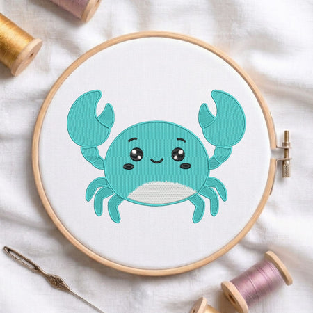A round embroidery hoop holding white fabric with a cute turquoise crab design stitched in the center. The hoop is styled on a white fabric background with spools of thread and a needle around it.
