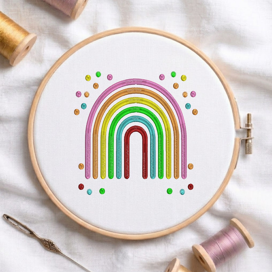 A circular embroidery hoop on white fabric, featuring a colorful rainbow design with glittery stripes and small rainbow dots. Spools of thread, a needle, and soft fabric are arranged around the hoop.