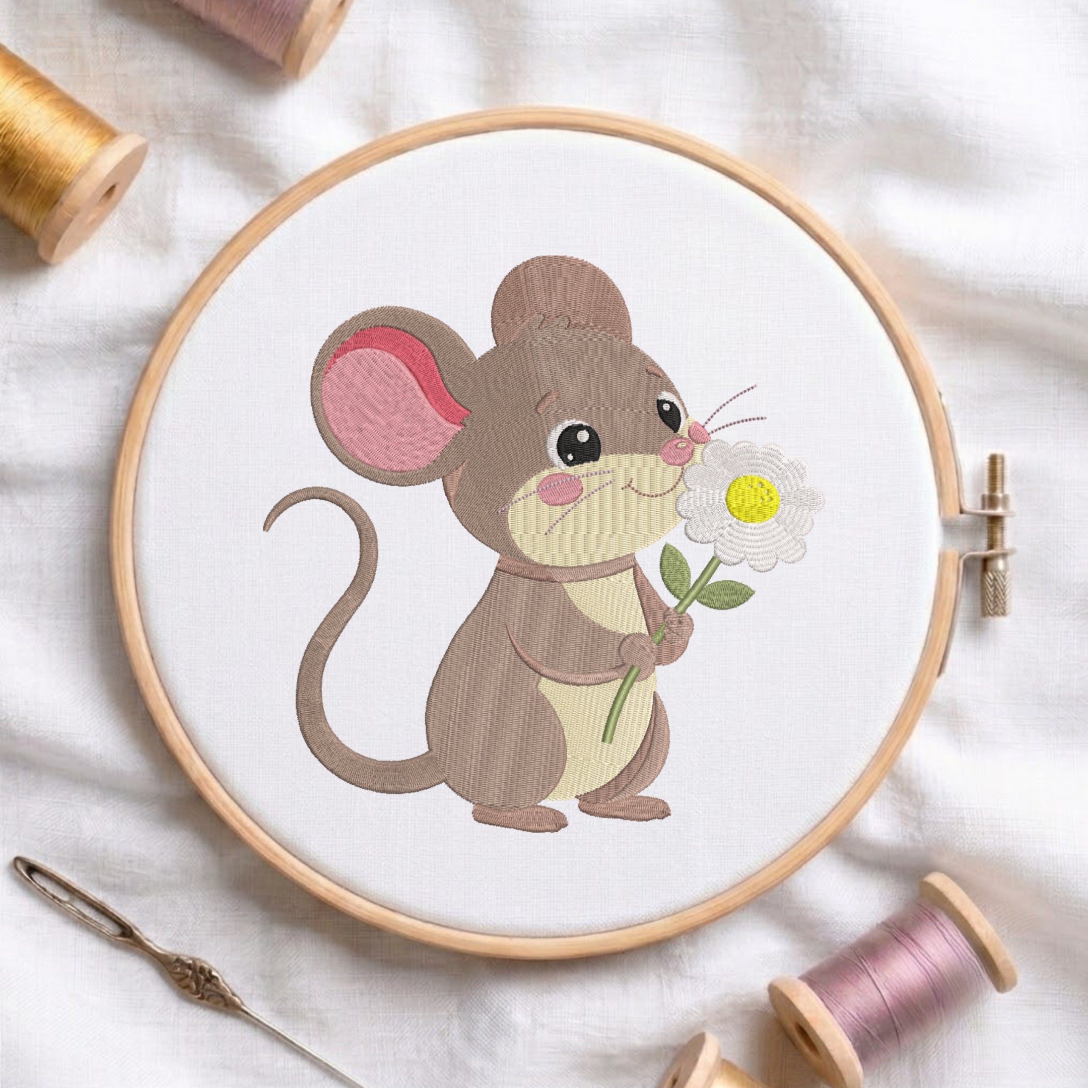 A circular embroidery hoop holding white fabric with a stitched design of a brown mouse holding a white flower with a yellow center. The hoop is styled on a white fabric background with spools of thread and a needle around it.