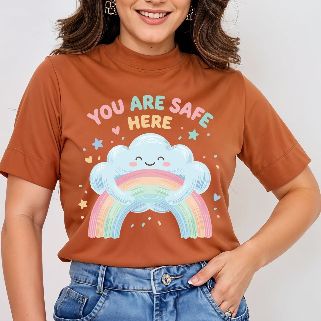 A woman wearing a rust-orange short-sleeve t-shirt with a colorful graphic of a smiling cloud holding a pastel rainbow and the text 'YOU ARE SAFE HERE'.