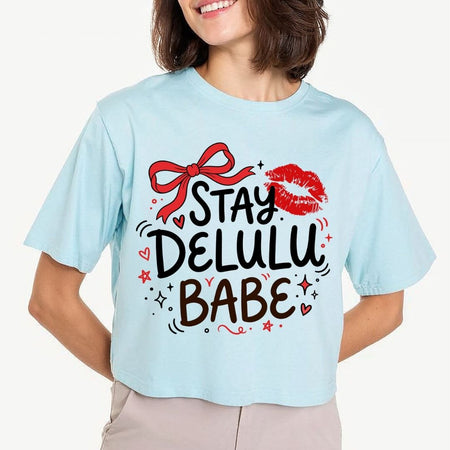 A woman wearing a light blue cropped t-shirt with a graphic that reads 'STAY DELULU BABE' in black and brown lettering, decorated with a red bow, red lips, and small red hearts, against a light gray background.
