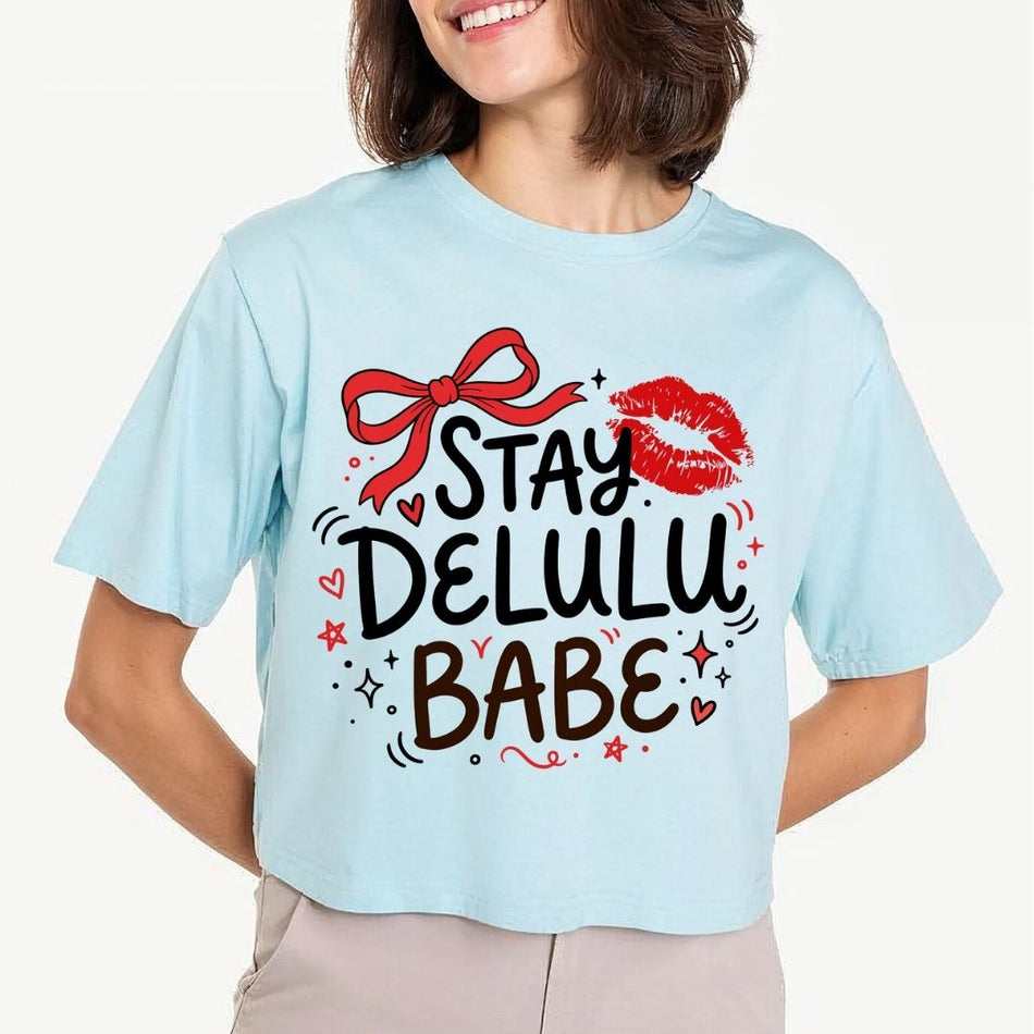 A woman wearing a light blue cropped t-shirt with a graphic that reads 'STAY DELULU BABE' in black and brown lettering, decorated with a red bow, red lips, and small red hearts, against a light gray background.