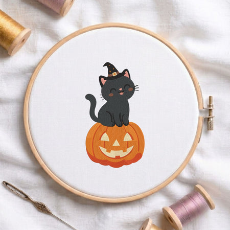 Cute black cat wearing a witch hat sitting on a jack-o-lantern pumpkin Halloween machine embroidery design shown in a wooden embroidery hoop on white fabric with thread spools and a needle