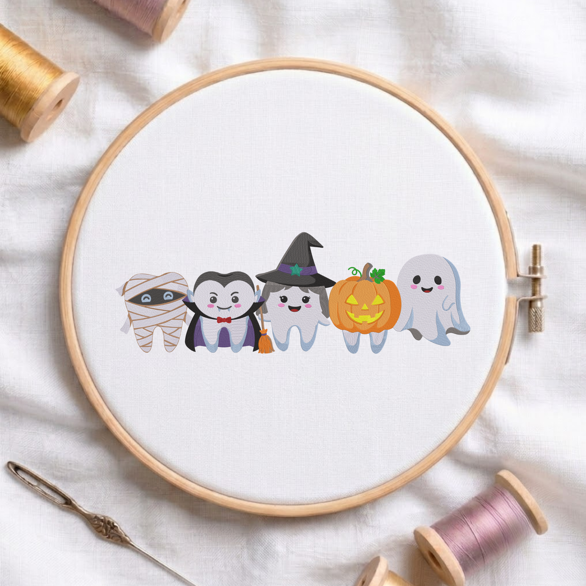 Cute Halloween dental teeth characters dressed as a mummy, vampire, witch, pumpkin, and ghost machine embroidery design shown in a wooden embroidery hoop on white fabric with thread spools and a needle