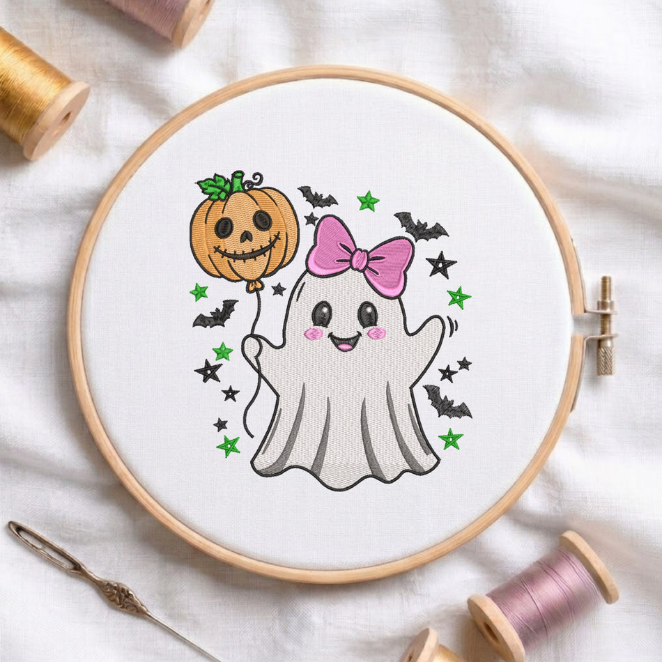 A circular embroidery hoop holding white fabric with a cute Halloween design: a white ghost with a pink bow, a smiling orange pumpkin, black bats, and green stars. The hoop is styled on a white fabric background with spools of thread and a needle around it.
