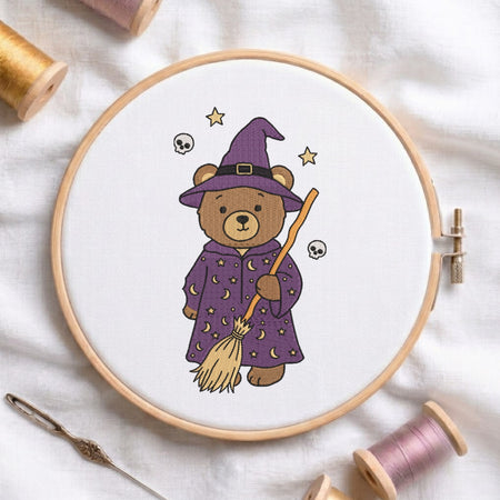 A circular embroidery hoop holds white fabric with a stitched design of a teddy bear dressed as a witch in a purple robe and hat, holding a broomstick. The hoop is styled on a white fabric background with spools of thread and a needle around it.
